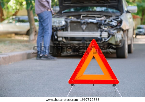 Red triangle warning sign of\
car accident on road in front of wrecked car. Man inspecting\
wrecked car after accident. Driver look under the hood of wrecked\
auto.