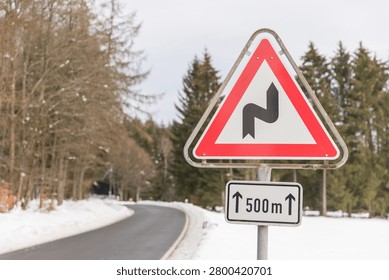 A red triangular double-bend warning sign with a 500m distance plate stands beside a snow-covered winding road through a winter forest of mixed bare and conifer trees.