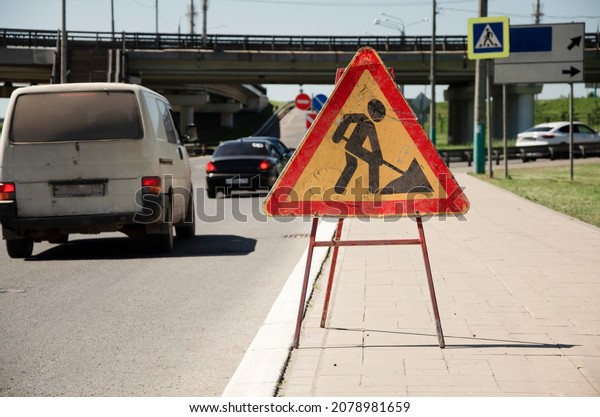 A red\
triangular sign near the road informs about road works. Road sign,\
repair work near the roadway in the\
city.