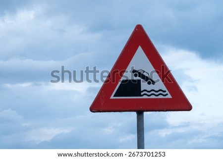 Red triangular warning road sign indicating danger of car falling into water, set against cloudy sky
