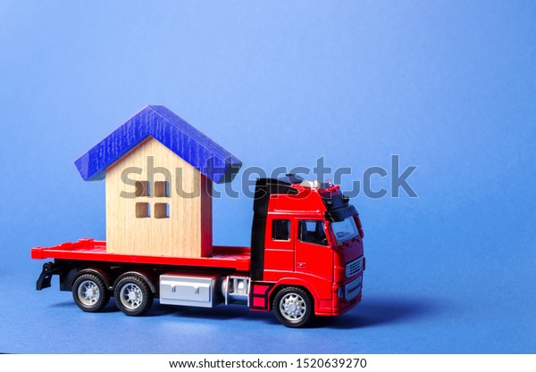 Red truck carrier transports a blue roofed\
house. Concept of transportation and cargo shipping, moving\
company. Construction of new houses and objects. Logistics and\
supply. Move entire\
buildings