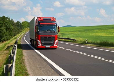 Red Truck Driving On Asphalt Road In A Rural Landscape. Green Fields And Alleys Leading Along The Road. Sunny Summer Day With A Cloudy Sky.