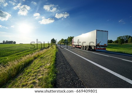 Red truck driving on asphalt road along the green fields in rural landscape at sunset