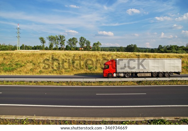 Red truck\
driving on asphalt highway beneath the grassy slope. Electricity\
pylons, forest and village houses on the horizon. Sunny summer day\
with blue skies and white\
clouds.