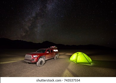 A Red Truck And Green Camping Tent Glow From Lights Inside. Set Up On A Flat Dry Lake Bed With The Night Sky And Milky Way Galaxy Overhead.