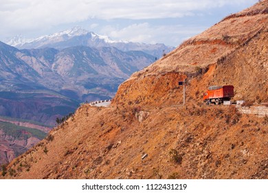 Red Truck Moving Slowly On The Red Mountain Road Through The Western Yunnan, China. Remote Area. Snow Mountains Background. Winter Season. Transportation Concept.