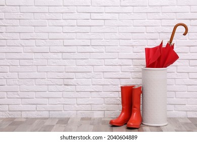 Red Umbrella In Holder And Rubber Boots Near White Brick Wall Indoors. Space For Text