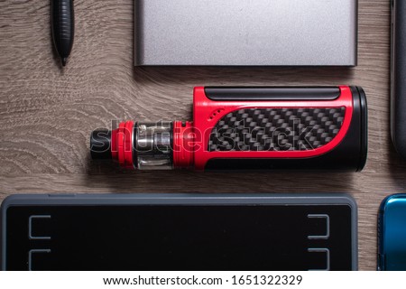 Red vaping device flat on a table surrounded by a graphics tablet and a drawing black pen. SIlver power bank