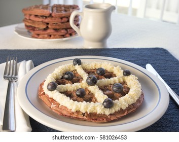 A Red Velvet Belgian Waffle Topped With Sweet Topping And Blueberries, With A Pitcher Of Maple Syrup And Larger Stack Of Waffles In The Background