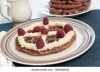 A Red Velvet Belgian Waffle Topped With Sweet Topping And Red Raspberries, With A Pitcher Of Maple Syrup And Larger Stack Of Waffles In The Background