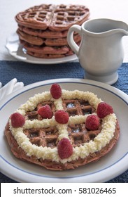 A Red Velvet Belgian Waffle Topped With Sweet Topping And Raspberries, With A Pitcher Of Maple Syrup And Larger Stack Of Waffles In The Background