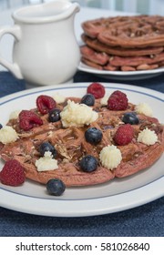 A Red Velvet Belgian Waffle Topped With Sweet Topping And Berries, With A Pitcher Of Maple Syrup And A Larger Stack Of Waffles In The Background