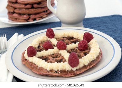 A Red Velvet Belgian Waffle Topped With Sweet Topping And Raspberries, With A Pitcher Of Maple Syrup And Larger Stack Of Waffles In The Background