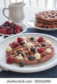 A Red Velvet Belgian Waffle Topped With Sweet Topping And Berries, With A Pitcher Of Maple Syrup, Larger Stack Of Waffles, And Bowl Of Mixed Berries In The Background