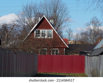 Red Vermillion Wooden House In The Spring Among The Fences In The Village And Small Town. Life In Russia Outside The Cities, Triangular Roof, Pre-war Buildings. Resettlement Of Dilapidated Housing