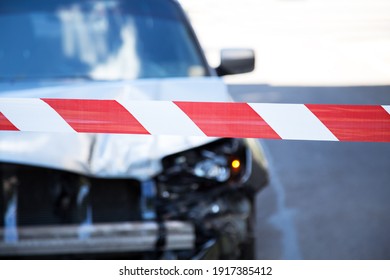 Red Warning Police Tape Afore Destroyed Car In Car Crash Traffic Accident On City Road. Smashed Broken Front Auto Headlight, Dented Hood Without Bumper On Gray Car Accident