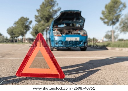 Red warning triangle placed on asphalt near a broken car with open hood, signaling vehicle breakdown