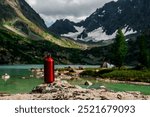 A red water bottle stands on a stone against the background of a mountain lake