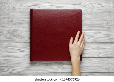 Red Wedding Photo Book With Leather Cover. Stylish Wedding Photo Album Close Up. Person Opens A Square Photobook. Family Burgundy Photoalbum On Wooden Background. Womans Hand Holding A  Photo Album