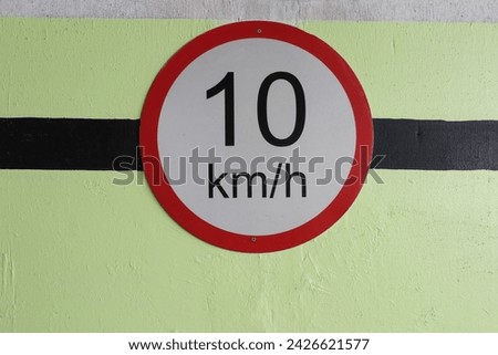 a red and white 10 km per hour speed control limit sign hanging on the wall in a carpark with green and black painted wall