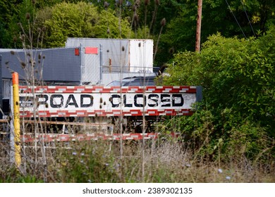 a red and white and black sign stating Road Closed surrounded by overgrown weeds and bushes with semi-trucks in the background