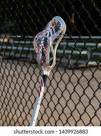 Red, White, And Blue Mens Lacrosse Stick Leaning Against A Chain Link Fence.