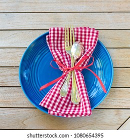 Red White And Blue Picnic Table Place Setting With Napkin, Fork, Spoon And Plate.  Square And Flat Layout Style Photo Taken Outside With Reflections Of Trees And Warm Sunlight In The Silverware.