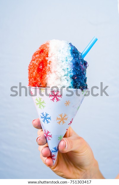 Red, white and blue snow cone in paper cone\
and blue straw held by female\
hands.