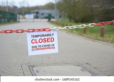 Red white chain barrier and sign with text Temporarily Closed due to Covid-19 Coronavirus, in front of a blurred company, countrywide pandemic lock down, copy space, selected focus