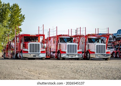 Red and white classic industrial pro big rig day cab semi trucks tractors with hydraulic modular two level car hauler semi trailers standing on the warehouse parking lot waiting for next freight