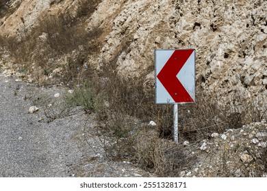 A red and white curved road warning sign stands beside a gravel road amidst rocky surroundings in rural Iran, indicating a sharp bend ahead. - Powered by Shutterstock