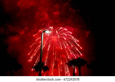 Red and white fireworks exploding in with palm trees in the background and red smoke in the night sky