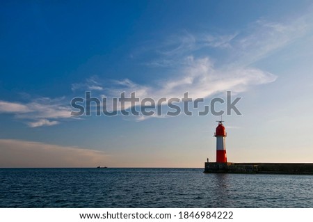 Red and white lighthouse and the calm Black Sea with an evening sunset sky. A small silhouette of a fisherman standing on the edge of the pier and a ship on the horizon. Summer seascape. Sochi, Russia