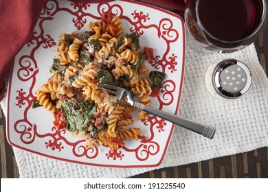 A Red And White Plate Filled With Fusilli Pasta, Prepared With Fresh Baby Spinach, Diced Tomatoes And Parmesan Cheese; A Glass Of Red Wine And Shaker Of Parmesan Cheese Sit Off To The Side