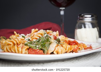 A Red And White Plate Filled With Fusilli Pasta, Prepared With Fresh Baby Spinach, Diced Tomatoes And Parmesan Cheese; A Glass Of Red Wine And Shaker Of Parmesan Cheese Sit Off To The Side