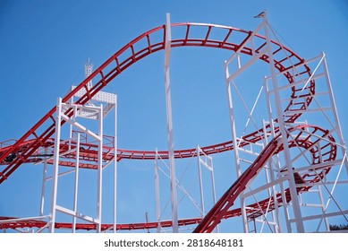 Red and white roller coaster loop at an amusement park under a clear blue sky with seagulls flying. Exciting summer attraction and fun concept for travel, family vacation, and thrill seekers.