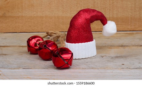 Red And White Santa Hat Next To Three Jingle Bells On A Wooden Background With Writing Space