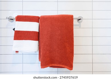 Red and white striped towel with a solid orange towel hanging on a chrome rack against a white tiled wall in a modern bathroom.