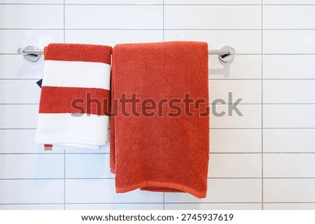 Red and white striped towel with a solid orange towel hanging on a chrome rack against a white tiled wall in a modern bathroom.