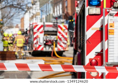 Red and white striped warning tape prevents people entering an incident in a town centre. In the distance, and intentionally out of focus, fire fighters in uniform, fire appliances and yellow hoses.