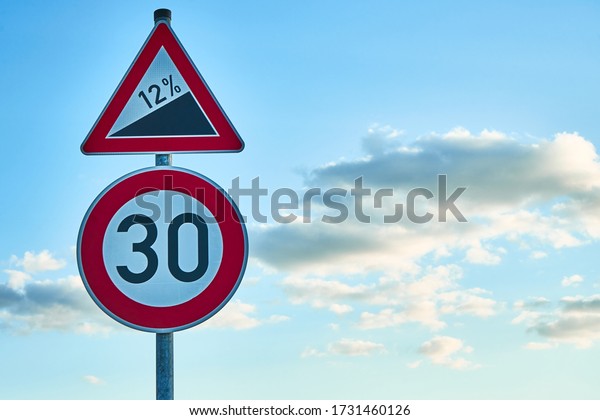 A red\
and white traffic sign, which warns of a twelve percent gradient\
and a traffic sign below it with a speed limit of 30 km/h. A blue\
sky with white clouds is in the\
background.