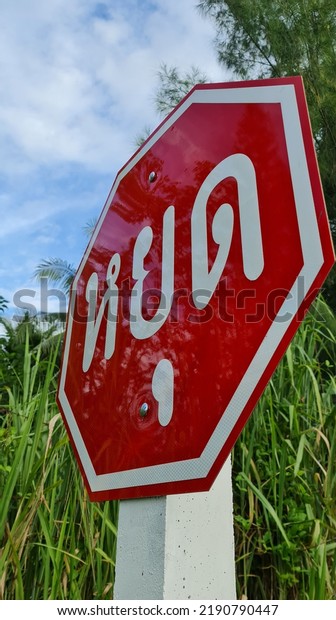 A red and white traffic sign with the word Stop\
on a white pole located at a junction blocked by trees in rural\
Thailand.