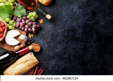 Red And White Wine Bottles, Grape, Cheese And Sausages Over Stone Table. Top View With Copy Space