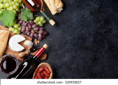 Red And White Wine Bottles, Grape, Cheese And Sausages Over Stone Table. Top View With Copy Space