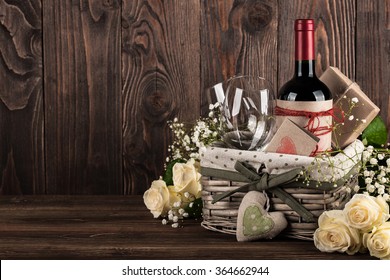 Red Wine Bottle, Two Wine Glasses, Gift Boxes In The Basket, White Roses On The Dark Wooden Background
