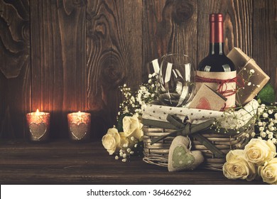 Red Wine Bottle, Two Wine Glasses, Gift Boxes In The Basket, White Roses On The Dark Wooden Background