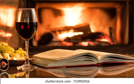 Red wine glass with an open book on table in front of burning fireplace.