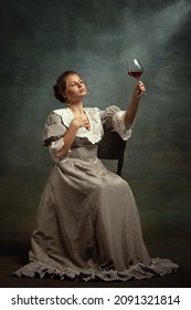 With red wine glass. Young beautiful girl in gray dress of medieval style sitting on chair isolated on dark background. Comparison of eras concept, flemish style. Classic art character, old-fashioned.