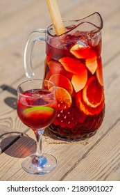 Red wine sangria decanter with apple, orange, lemon, lime wedges, raspberries, grapes and ice. There is a glass of ice, raspberries and sangria nearby. Stands on a wooden background illuminated by sun