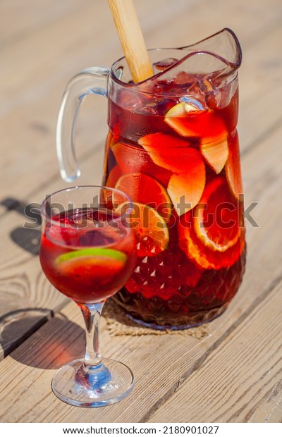 Red wine sangria decanter with apple, orange, lemon,\
lime wedges, raspberries, grapes and ice. There is a glass of ice,\
raspberries and sangria nearby. Stands on a wooden background\
illuminated by sun